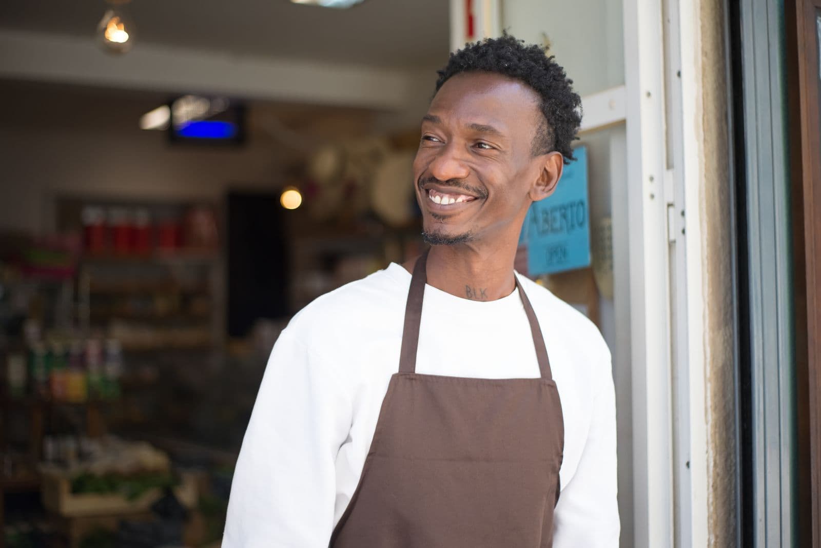 A smiling shop owner in an apron standing at the entrance of his store